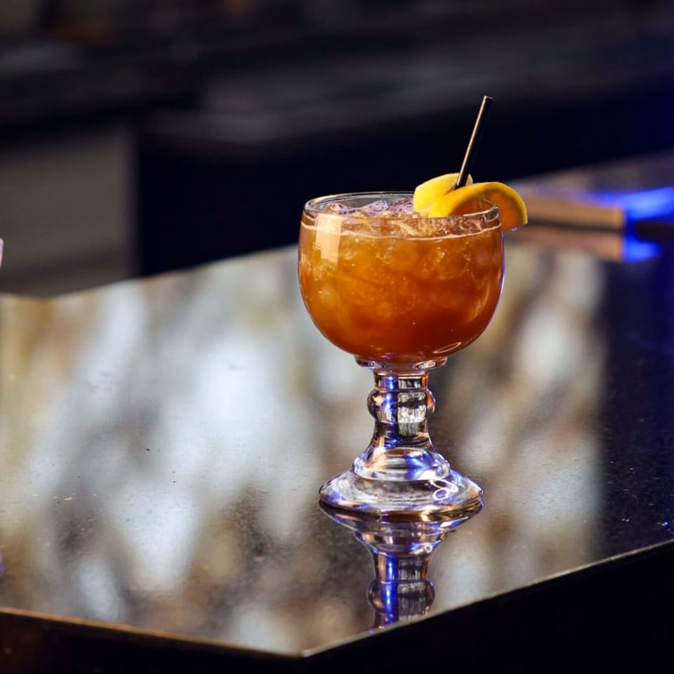 A glass of Encanto Alebrije's Mexican cocktail with ice and an orange slice on a bar.