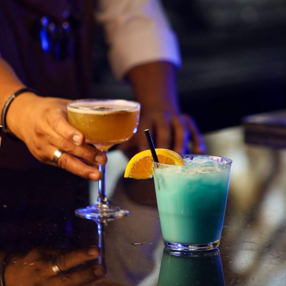 Bartender's hands serving a golden cocktail and a blue cocktail with an orange slice.
