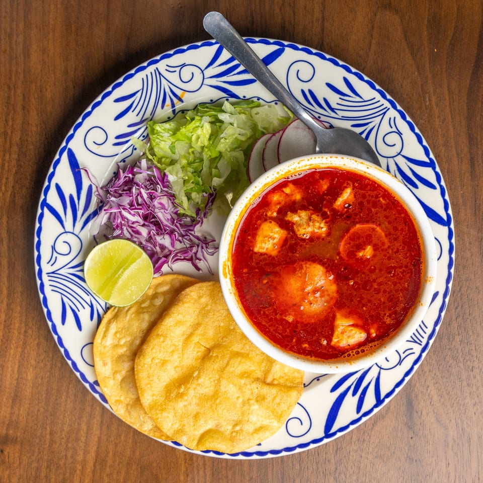 Encanto Alebrije's red pozole soup with tortillas, lime, lettuce, and cabbage on a decorative plate.
