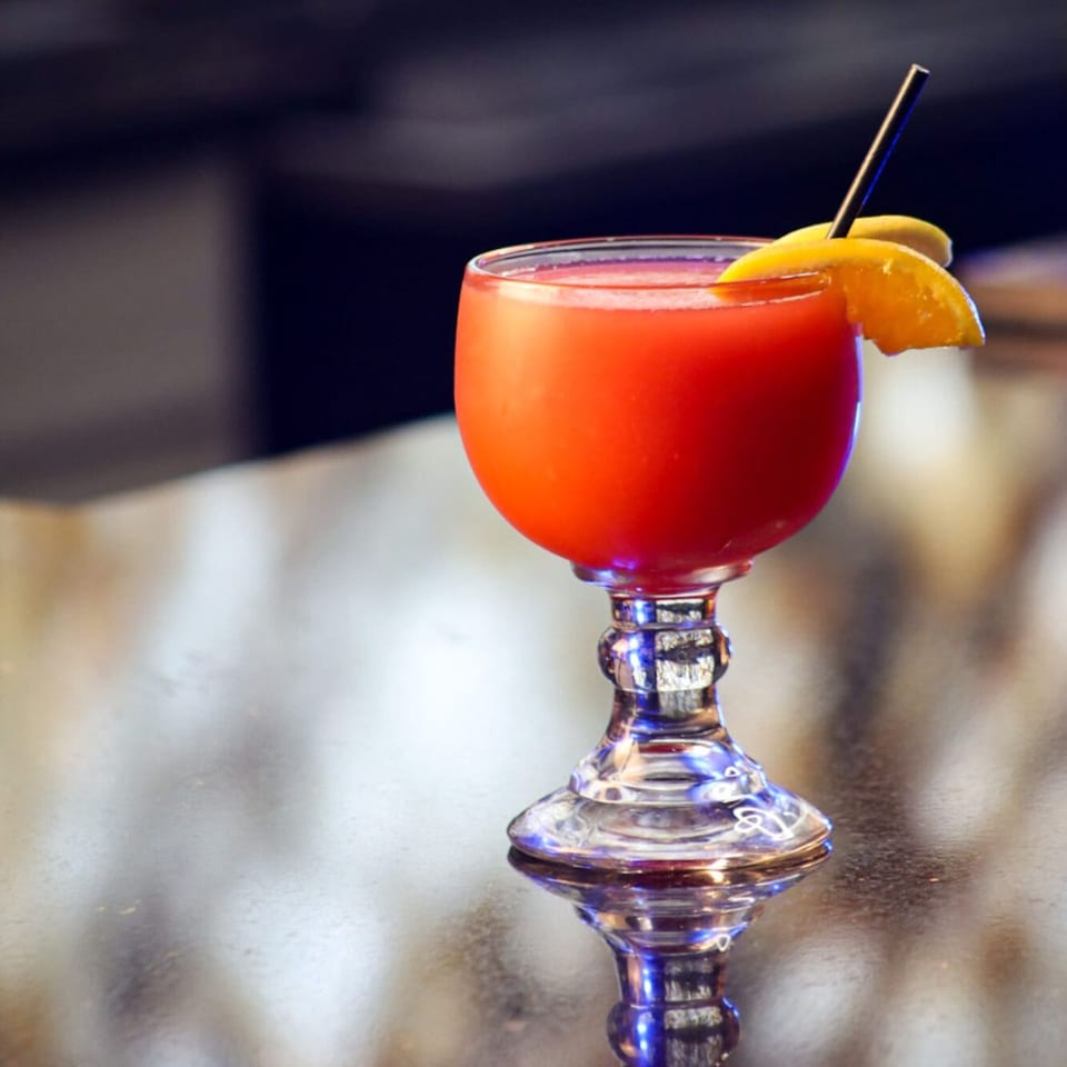 A vibrant red cocktail in a decorative glass, garnished with an orange slice and a black straw.