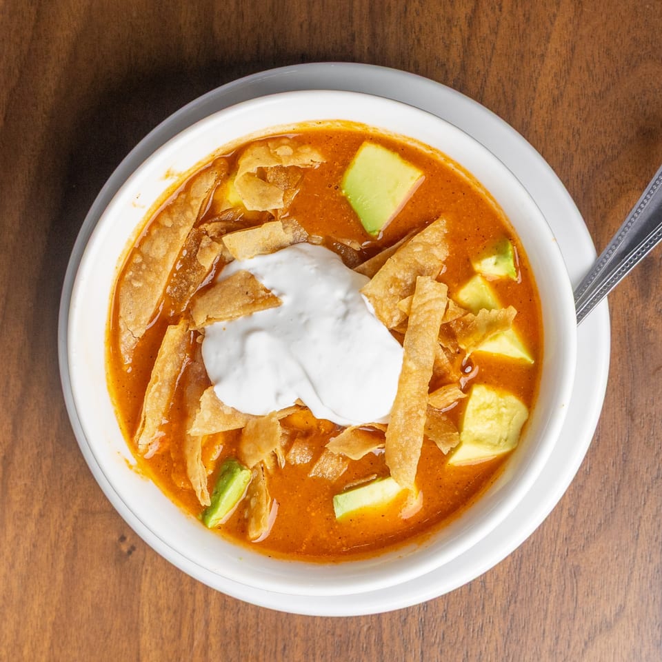 Encanto Alebrije's tortilla soup with avocado, tortilla strips, and sour cream in a white bowl.
